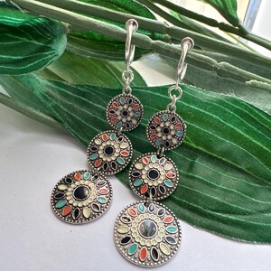 May include: A pair of silver dangle earrings with three round, colorful, enamel-painted charms. The charms feature a black center with a floral design in shades of white, orange, and blue.