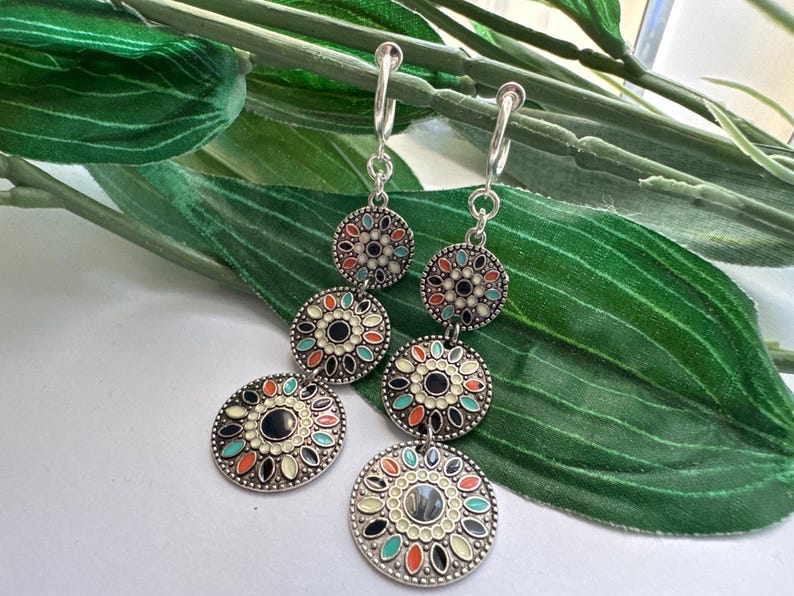 May include: A pair of silver dangle earrings with three round, colorful, enamel-painted charms. The charms feature a black center with a floral design in shades of white, orange, and blue.