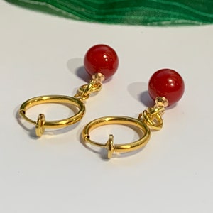 Puede incluir: Un par de pendientes de clip con un dise&ntilde;o de aro dorado y un peque&ntilde;o cierre. Cada pendiente tiene una cuenta redonda de color rojo intenso suspendida de un nudo dorado. Los pendientes se muestran sobre una superficie blanca.
