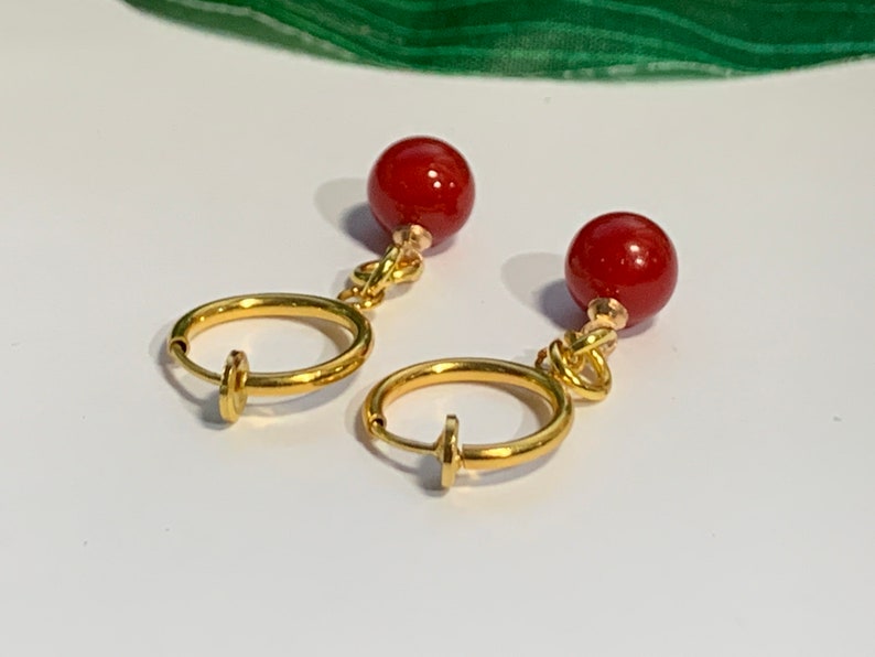 Puede incluir: Un par de pendientes de clip con un dise&ntilde;o de aro dorado y un peque&ntilde;o cierre. Cada pendiente tiene una cuenta redonda de color rojo intenso suspendida de un nudo dorado. Los pendientes se muestran sobre una superficie blanca.