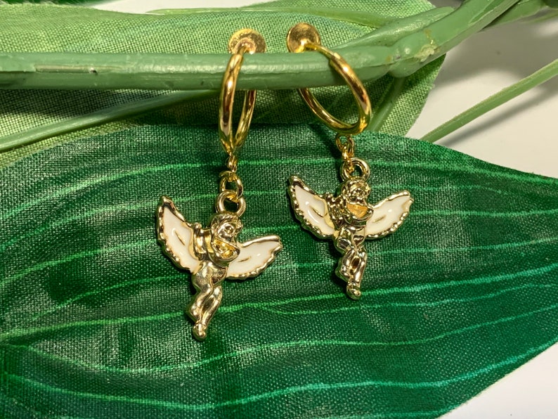 May include: A pair of gold hoop earrings with angel charms. The angels are white with gold accents and have wings.