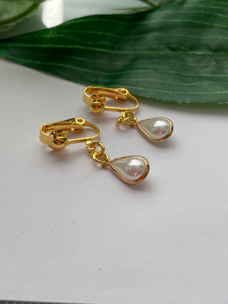 May include: A pair of gold-toned clip-on earrings with a teardrop-shaped, faux pearl pendant. The earrings have a classic design, with a shiny finish. The background is a soft white surface with a green leaf.