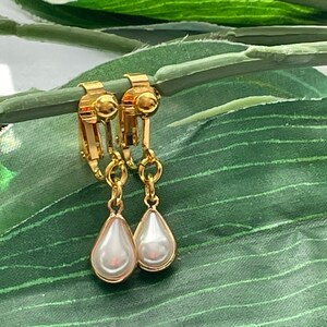 May include: A pair of gold-toned clip-on earrings with pear-shaped, faux pearl drops. The earrings are displayed on a green leaf, showcasing their elegant design and classic appeal. The clip-on style offers a comfortable fit.