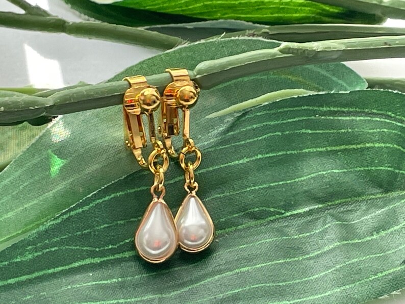 May include: A pair of gold-toned clip-on earrings with pear-shaped, faux pearl drops. The earrings are displayed on a green leaf, showcasing their elegant design and classic appeal. The clip-on style offers a comfortable fit.