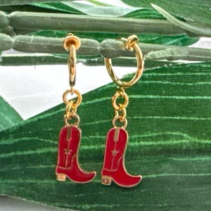 May include: Gold-colored hoop earrings with dangling red cowboy boot charms. The boots have white star details and a gold heel. The earrings are set against a backdrop of green leaves.