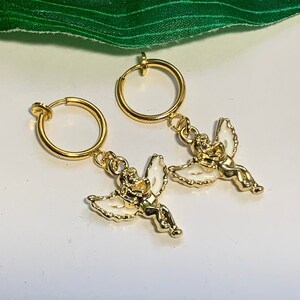 May include: A pair of gold clip-on earrings with a small angel charm hanging from each hoop.