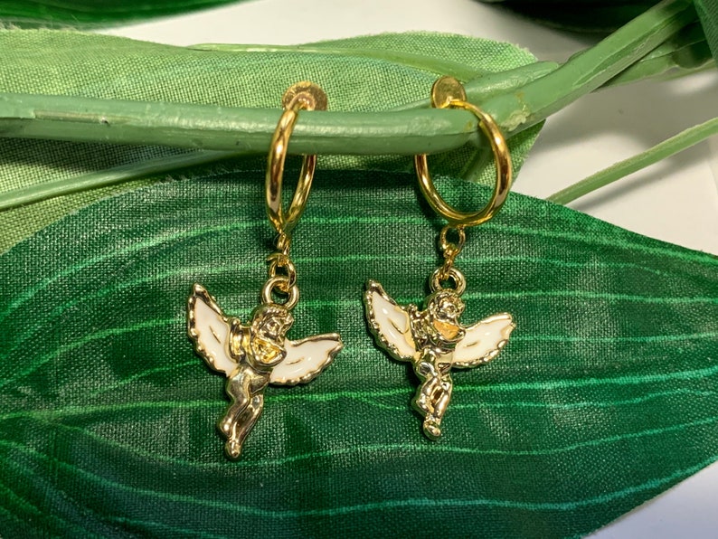 May include: A pair of gold hoop earrings with a white enamel angel charm hanging from each hoop. The angels are holding a heart in their hands.