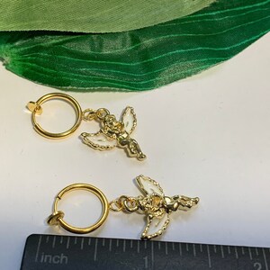 May include: Two gold-toned clip-on earrings with a white angel charm. Each earring has a circular hoop and a small angel charm with white wings and a gold-toned body.