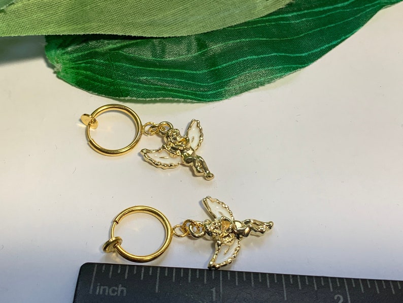 May include: Two gold-toned clip-on earrings with a white angel charm. Each earring has a circular hoop and a small angel charm with white wings and a gold-toned body.