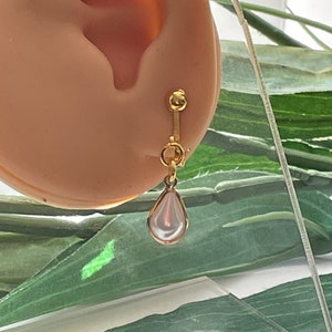 May include: A gold-colored clip-on earring with a pear-shaped, white, pearl-like drop. The earring is attached to a light-colored ear. The background features green leaves and a clear surface.