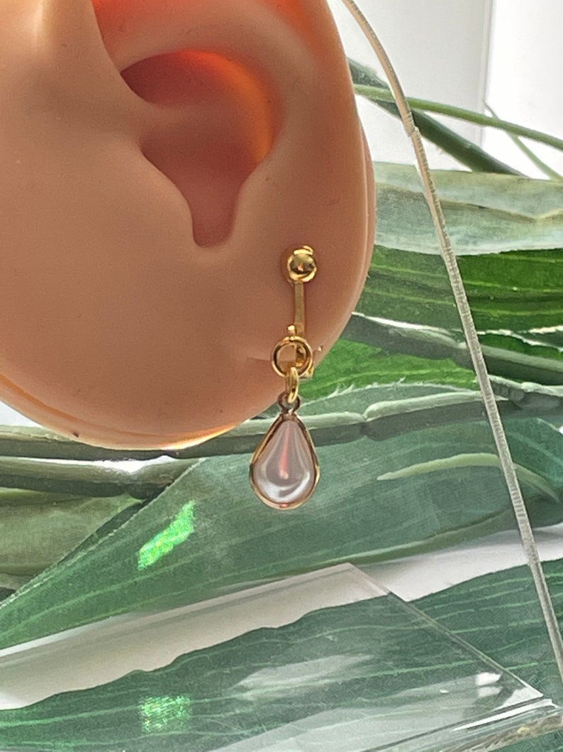 May include: A gold-colored clip-on earring with a pear-shaped, white, pearl-like drop. The earring is attached to a light-colored ear. The background features green leaves and a clear surface.