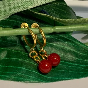 Puede incluir: Pendientes de aro dorados, cada uno con una peque&ntilde;a cuenta redonda roja colgando. Los pendientes se muestran sobre un fondo verde, resaltando el contraste entre el oro y el rojo.