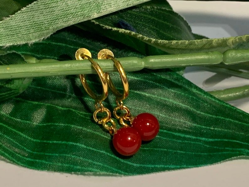 Puede incluir: Pendientes de aro dorados, cada uno con una peque&ntilde;a cuenta redonda roja colgando. Los pendientes se muestran sobre un fondo verde, resaltando el contraste entre el oro y el rojo.