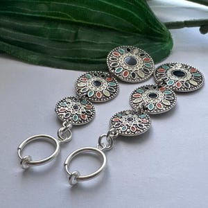 May include: A pair of silver hoop earrings with three decorative round charms. The charms are silver with a white background and feature a colorful floral design with green, red, and blue accents.