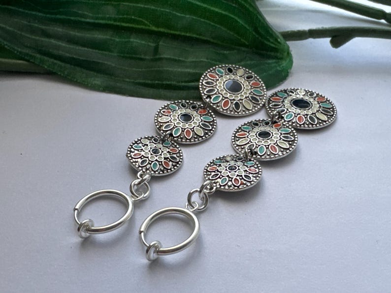 May include: A pair of silver hoop earrings with three decorative round charms. The charms are silver with a white background and feature a colorful floral design with green, red, and blue accents.