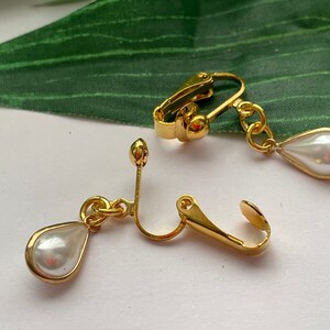 May include: Gold-toned clip-on earrings featuring a pear-shaped, faux pearl pendant. The earrings have a classic design with a gold frame around the pearl. The clip-on mechanism is visible, showing the earring's secure attachment style. These earrings are suitable for various occasions.