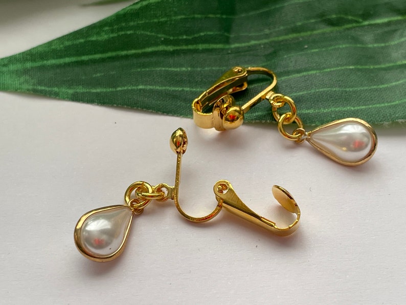 May include: Gold-toned clip-on earrings featuring a pear-shaped, faux pearl pendant. The earrings have a classic design with a gold frame around the pearl. The clip-on mechanism is visible, showing the earring's secure attachment style. These earrings are suitable for various occasions.