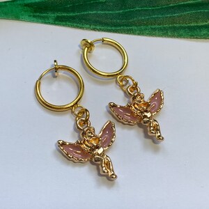 May include: A pair of gold hoop earrings with a pink angel charm dangling from each hoop.