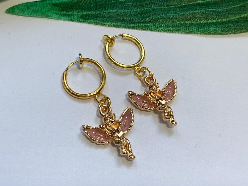 May include: A pair of gold hoop earrings with a pink angel charm dangling from each hoop.