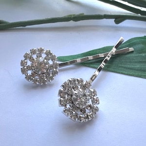 May include: Two silver-toned hair grips, each featuring a circular decorative element at the end. The circular ornaments are encrusted with numerous small, clear, faceted rhinestones arranged in a floral pattern around a larger central stone.