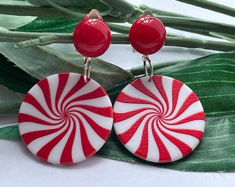 May include: A pair of clip-on earrings featuring a red and white peppermint swirl design. Each earring has a red, round top and a circular pendant with a spiral pattern. The earrings are set against a backdrop of green leaves.