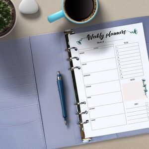 Könnte beinhalten: Ein lilafarbener Planer mit einer geöffneten weißen Wochenplanseite. Die Seite hat ein florales Design und den Text "Weekly Planners" und "Week of".
