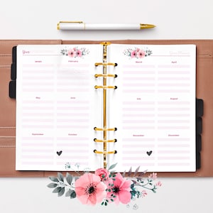 Puede incluir: Una agenda blanca con un diseño floral en la portada. La agenda está abierta a una vista de calendario anual con los meses etiquetados de enero a diciembre. La agenda tiene una encuadernación de anillos dorados.