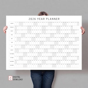 May include: A large white 2026 Year Planner with a grid layout, held by a person. The planner features the year's months and dates, with a 'Digital Download' icon in the bottom left corner. The text is in black.