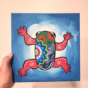 May include: A colorful painting of a frog on a blue canvas. The frog is painted in a folk art style with bright colors and intricate patterns. The frog's body is green, red, yellow, and blue, and its legs are pink. The background is a light blue.