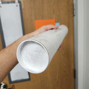 May include: A white mailing tube with a silver cap, wrapped in clear plastic, is being held by a hand.