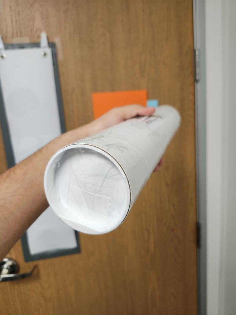 May include: A white mailing tube with a silver cap, wrapped in clear plastic, is being held by a hand.