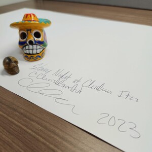 May include: A yellow and blue painted sugar skull wearing a colourful sombrero. The skull is sitting on a white piece of paper with the text "Snowy Night at Clarendon 1722 2023" written in black ink.