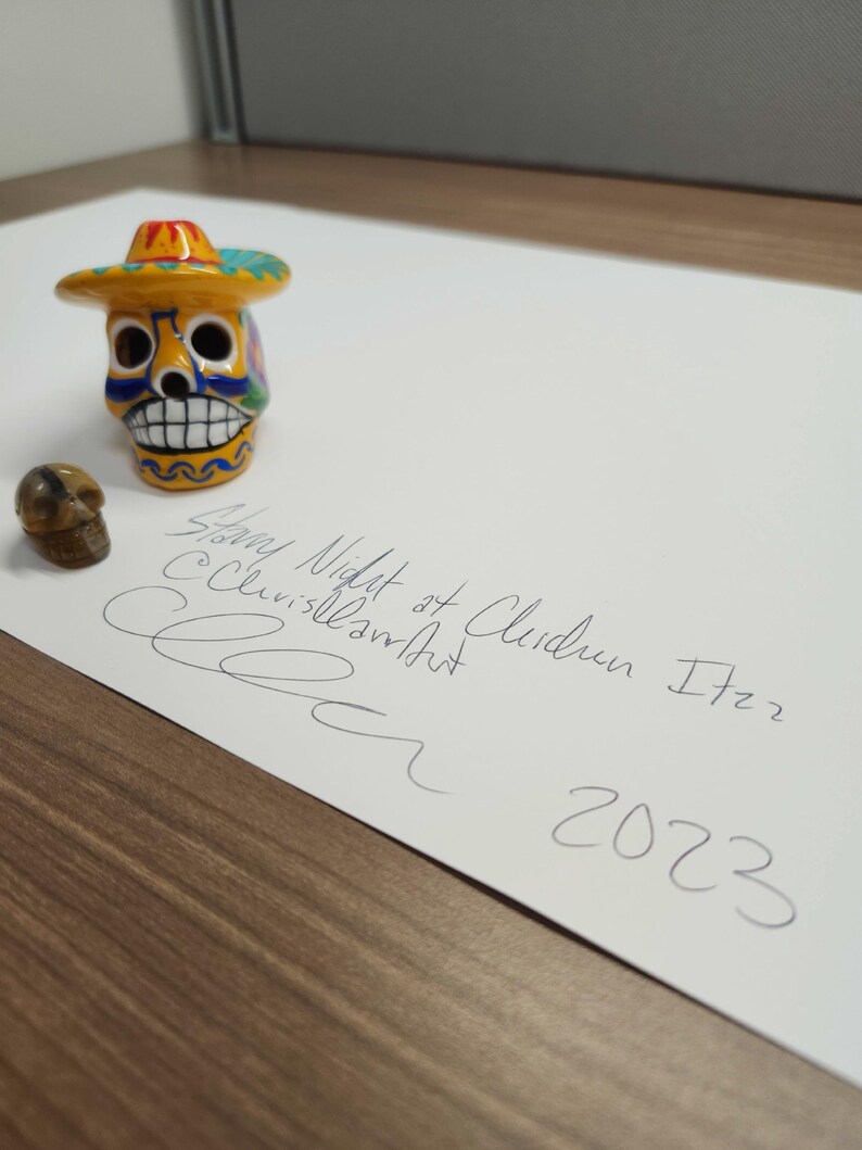 May include: A yellow and blue painted sugar skull wearing a colourful sombrero. The skull is sitting on a white piece of paper with the text "Snowy Night at Clarendon 1722 2023" written in black ink.