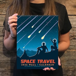 May include: A 2026 wall calendar with a space travel theme. The artwork depicts two silhouetted figures on a mountain, gazing at shooting stars against a dark blue starry sky. The calendar has the text "SPACE TRAVEL" and "ARTWORK BY ANDERSON DESIGN GROUP."