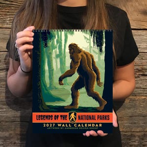 May include: A 2027 wall calendar featuring a brown Bigfoot illustration in a green forest setting. The text reads Legends of the National Parks, 2027 Wall Calendar, and Artwork by Anderson Design Group, with a small Bigfoot icon in the center.