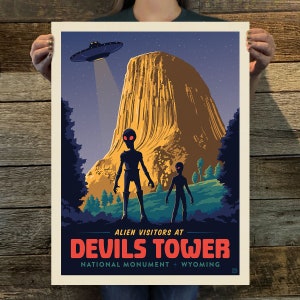 May include: A retro-style travel poster featuring a dark silhouette of two aliens standing in front of Devils Tower National Monument in Wyoming. The poster has a dark blue background with a flying saucer above the tower. The text reads "Alien Visitors at Devils Tower National Monument + Wyoming".