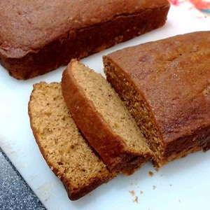 May include: Two loaves of freshly baked brown bread, one sliced into three pieces, on a white cutting board.