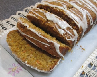 May include: A loaf of carrot cake with white icing is sliced and ready to be served. The cake is on a white surface with a crocheted doily underneath.
