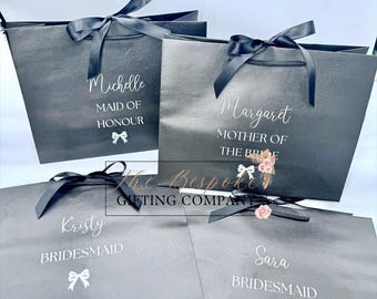 May include: Four matte black gift bags with black ribbon handles and bows. Each bag features white text with names including Michelle, Margaret, Kristy, and Sara, along with titles like Maid of Honour, Mother of the Bride, and Bridesmaid, plus a small bow icon.