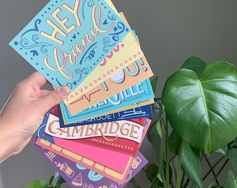 Puede incluir: Una pila de postales coloridas con varios diseños. La tarjeta superior es azul claro con el texto "Hey friend" en una fuente caprichosa. La siguiente tarjeta es amarilla con el texto "Love you!" en una fuente en negrita. La tercera tarjeta es azul con el texto "Cambridge, Massachusetts" en blanco. La cuarta tarjeta es rosa con una variedad de iconos coloridos.