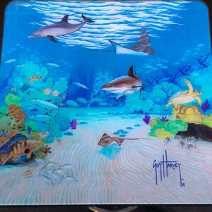 May include: A colourful underwater scene featuring a dolphin, a stingray, a shark, a turtle, and various fish. The scene is painted on a square glass table top with a signature by Guy Harvey.