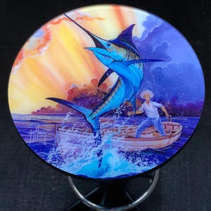 Puede incluir: Una mesa redonda de vidrio de colores con una pintura de un marlin azul saltando fuera del agua. Un hombre con camisa blanca y sombrero está en un pequeño bote de madera. El cielo es de un naranja y morado vibrante con nubes. La firma del artista, "Giff Harvey", está en la esquina inferior izquierda.