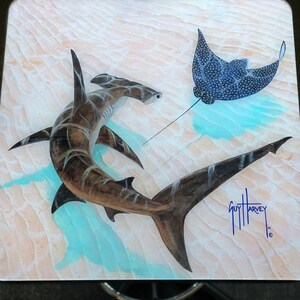 May include: A square wooden table top featuring a hammerhead shark and a spotted eagle ray swimming in a turquoise sea. The artwork is signed "Guy Harvey". The background has a textured, sandy appearance.
