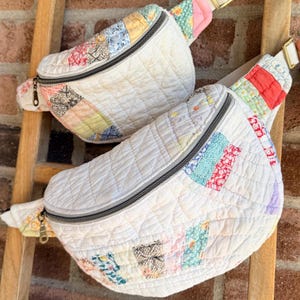 May include: Two crescent-shaped fanny packs constructed from quilted fabric featuring a patchwork design of white, yellow, blue, pink, and red floral and geometric patterns. Each bag includes a top zipper closure and an adjustable strap.