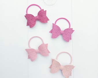 May include: Four pink hair ties with bows. The bows are made of textured fabric and are in different shades of pink.