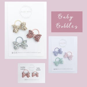 May include: Three sets of glitter hair bobbles in different colors. The first set is gold, the second is rose gold, and the third is silver. Each set has two bobbles. The text "Baby Bobbles" is written in pink letters on a pink background.