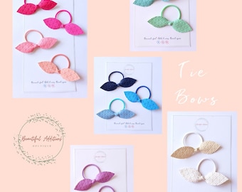 May include: Set of five pairs of tie bows in various colors, including pink, blue, green, and beige. Each bow is made of a textured fabric and is attached to a hair elastic.
