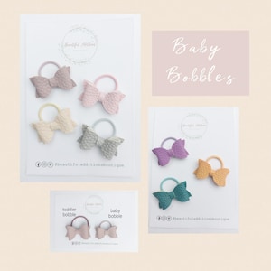 May include: Four sets of faux leather hair bows in various colors, including pink, beige, purple, teal, and yellow. The bows are arranged on white cards with the text "Baby Bobbles" and "Beautiful Additions Boutique".
