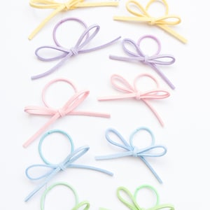 Può includere: Una collezione di fiocchi per capelli dai colori pastello, in taglie per neonati e bambini piccoli. I fiocchi sono nelle tonalità del giallo, lavanda, rosa, blu e verde. Il testo "Toddler size" e "Baby size" è visibile.