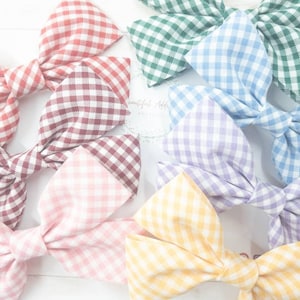 May include: A set of six gingham hair bows in various pastel colours, including pink, yellow, blue, green, and purple. The bows are made of a soft, textured fabric and are tied with a simple knot.